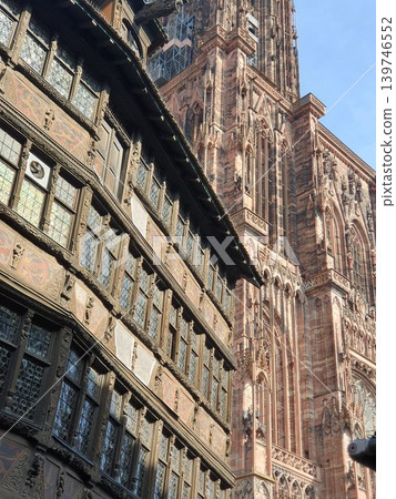 Sunlit Gothic cathedral towers beside a richly carved timber-framed building, showcasing medieval stonework, leaded windows and ornate decorative details. Urban heritage scene history, craftsmanship Sunlit Gothic cathedral towers beside a richly carved timber-framed building, showcasing medieval stonework, leaded windows and ornate decorative details. Urban heritage scene history, craftsmanship 139746552