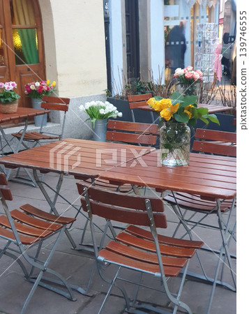 Bright floral arrangements on wooden bistro tables create a cozy outdoor cafe atmosphere in a charming courtyard, featuring roses, tulips and potted plants for a welcoming, romantic urban setting. Bright floral arrangements on wooden bistro tables create a cozy outdoor cafe atmosphere in a charming courtyard, featuring roses, tulips and potted plants for a welcoming, romantic urban setting. 139746555
