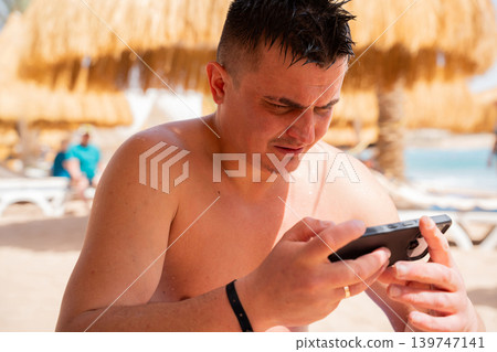 Caucasian man using smartphone at beach resort in Sharm El Sheikh Egypt near Red Sea umbrellas. Remote work and travel lifestyle Caucasian man using smartphone at beach resort in Sharm El Sheikh Egypt near Red Sea umbrellas. Remote work and travel lifestyle 139747141