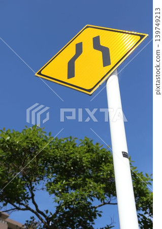 A yellow diamond-shaped sign indicates a reduction in road lanes. A yellow diamond-shaped sign indicates a reduction in road lanes. 139749213