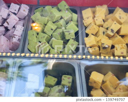 Colorful assortment of traditional Indian sweets displayed in a glass case, featuring pink, green, and yellow varieties with distinct shapes and textures Colorful assortment of traditional Indian sweets displayed in a glass case, featuring pink, green, and yellow varieties with distinct shapes and textures 139749707