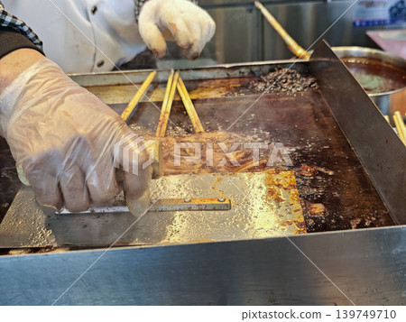 Chef in white gloves prepares skewered meat in a hot oil fryer at a street food stall, with cooking utensils and ingredients visible in the background Chef in white gloves prepares skewered meat in a hot oil fryer at a street food stall, with cooking utensils and ingredients visible in the background 139749710