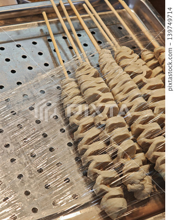 Raw skewered meat wrapped in plastic film resting on a perforated metal tray, ready for cooking in a kitchen setting Raw skewered meat wrapped in plastic film resting on a perforated metal tray, ready for cooking in a kitchen setting 139749714