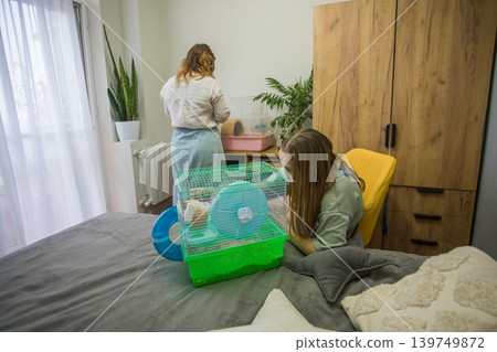 Two people are cleaning a hamster cage. One person is sitting on a bed  139749872
