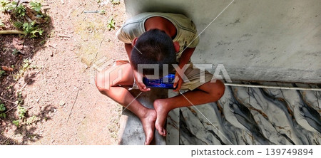 Serang, Banten, Indonesia, 05-18-2026, Boy sitting playing games on his phone on the porch Serang, Banten, Indonesia, 05-18-2026, Boy sitting playing games on his phone on the porch 139749894