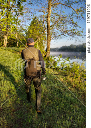 A person walks along the riverbank in the morning light, holding a fishing rod. Trees surround the area, and a gentle mist rises off the water A person walks along the riverbank in the morning light, holding a fishing rod. Trees surround the area, and a gentle mist rises off the water 139751908