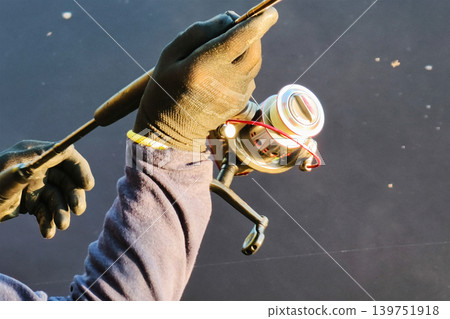 A person grips a fishing rod with a reel in hand by the river as the sun rises. The dark water reflects the light of dawn creating a still scene A person grips a fishing rod with a reel in hand by the river as the sun rises. The dark water reflects the light of dawn creating a still scene 139751918