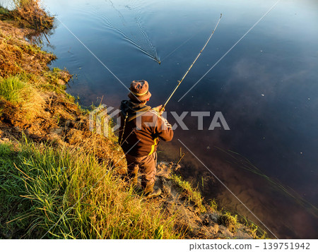A person stands on the bank of a river early in the morning, holding a fishing rod and waiting for a catch while surrounded by grass and calm waters A person stands on the bank of a river early in the morning, holding a fishing rod and waiting for a catch while surrounded by grass and calm waters 139751942