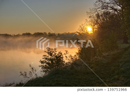 Sun rises above the river as mist rises from the water. Trees line the bank, and the sky shows warm colors in the early morning Sun rises above the river as mist rises from the water. Trees line the bank, and the sky shows warm colors in the early morning 139751966