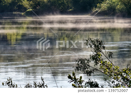 Mist hovers above the still water in the early morning as sunlight breaks through the trees along the green riverbank in a natural setting Mist hovers above the still water in the early morning as sunlight breaks through the trees along the green riverbank in a natural setting 139752007