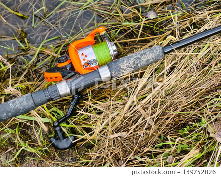 A fishing rod and reel rest on dry grass by a body of water. The bright reel contrasts with the earth tones around it. This scene shows a moment before fishing starts A fishing rod and reel rest on dry grass by a body of water. The bright reel contrasts with the earth tones around it. This scene shows a moment before fishing starts 139752026
