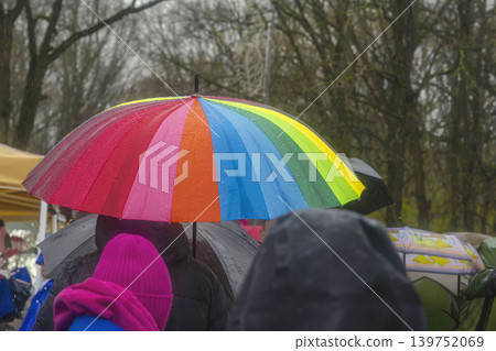 Rainbow umbrella covers people standing in crowded in rain, contrast sharply with overall gray, muted background, atmosphere of late autumn, early spring. Cozy melancholy of rainy weather concept Rainbow umbrella covers people standing in crowded in rain, contrast sharply with overall gray, muted background, atmosphere of late autumn, early spring. Cozy melancholy of rainy weather concept 139752069