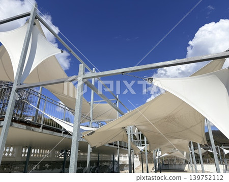 a modern outdoor shading structure featuring multiple white tensile fabric canopies stretched taut over a framework of galvanized steel beams and vertical posts. a modern outdoor shading structure featuring multiple white tensile fabric canopies stretched taut over a framework of galvanized steel beams and vertical posts. 139752112