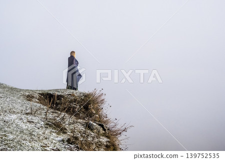 Solitary figure stands atop misty cliff edge wind tugs at dark garment 139752535