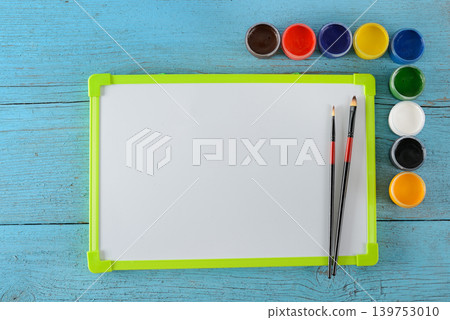 Creative workspace with white drawing board and set of vibrant paint containers on weathered blue timber texture 139753010