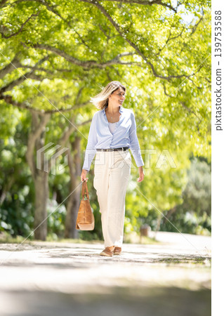 Woman walks along a path in a park under green trees during a sunny day in the afternoon Woman walks along a path in a park under green trees during a sunny day in the afternoon 139753588
