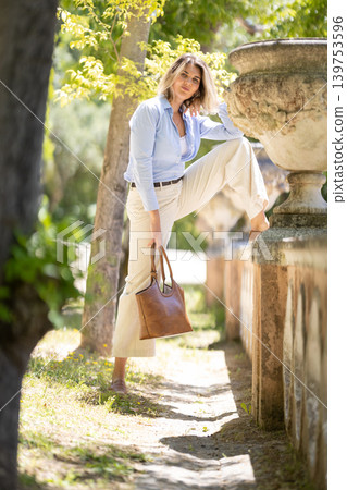 Woman poses with one leg raised on a stone wall while holding a brown bag in a garden during daytime Woman poses with one leg raised on a stone wall while holding a brown bag in a garden during daytime 139753596
