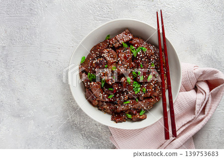 Crispy beef, with green onions and sesame seeds, in a bowl with chopsticks, on a textured surface, on a light background, no people Crispy beef, with green onions and sesame seeds, in a bowl with chopsticks, on a textured surface, on a light background, no people 139753683