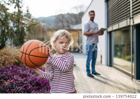 Toddler boy playing with basketball while father controls smart home system on terrace. 139754022
