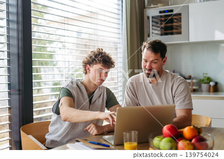 Father helping teenage son with diabetes with homework on laptop in kitchen. 139754024