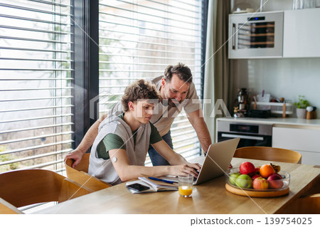 Father helping teenage son with diabetes with homework on laptop in kitchen. 139754025