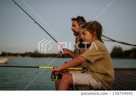 Father and son fishing together from wooden pier at sunset during family leisure time. Father and son fishing together from wooden pier at sunset during family leisure time. 139754045