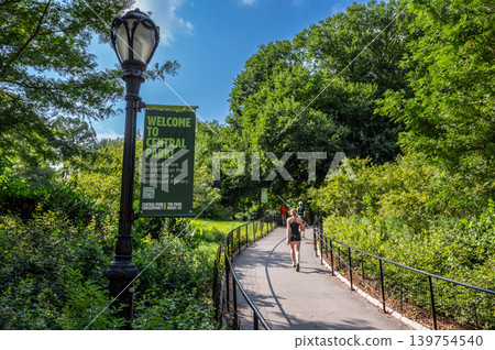 New york, usa august 18,2024. Central park welcome sign on lamppost with joggers and walkers on a sunny, tree lined manhattan pathway in new york city New york, usa august 18,2024. Central park welcome sign on lamppost with joggers and walkers on a sunny, tree lined manhattan pathway in new york city 139754540