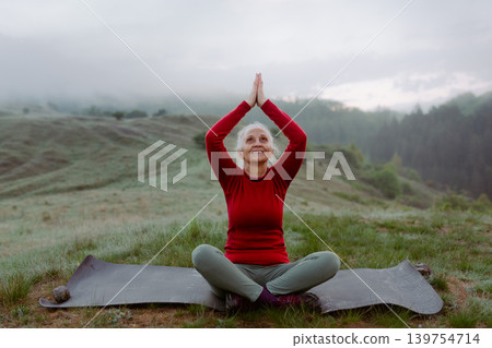 Senior woman meditating in nature on early morning with fog and mountains in background. Senior woman meditating in nature on early morning with fog and mountains in background. 139754714