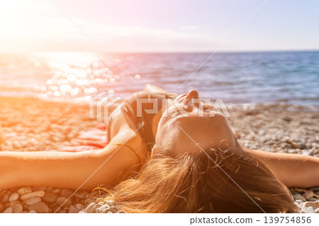 Woman beach sunbathing while lying on a pebble beach under the warm summer sun with the blue ocean in the background Woman beach sunbathing while lying on a pebble beach under the warm summer sun with the blue ocean in the background 139754856
