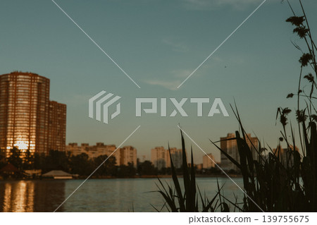 City lake at golden hour with reeds in foreground and modern buildings reflection. Peaceful urban nature landscape, sunset light on water, and residential area skyline. Calm summer evening vibe. City lake at golden hour with reeds in foreground and modern buildings reflection. Peaceful urban nature landscape, sunset light on water, and residential area skyline. Calm summer evening vibe. 139755675