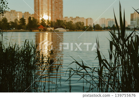 City lake at golden hour with reeds in foreground and modern buildings reflection. Peaceful urban nature landscape, sunset light on water, and residential area skyline. Calm summer evening vibe. City lake at golden hour with reeds in foreground and modern buildings reflection. Peaceful urban nature landscape, sunset light on water, and residential area skyline. Calm summer evening vibe. 139755676