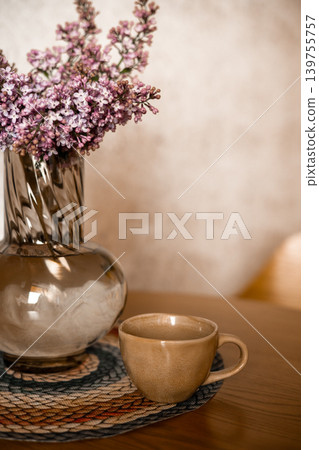 Spring lilac flowers in elegant glass vase next to ceramic coffee cup on wooden table. Cozy home morning atmosphere, warm neutral tones, slow living concept, and soft sunlight aesthetic. Spring lilac flowers in elegant glass vase next to ceramic coffee cup on wooden table. Cozy home morning atmosphere, warm neutral tones, slow living concept, and soft sunlight aesthetic. 139755757