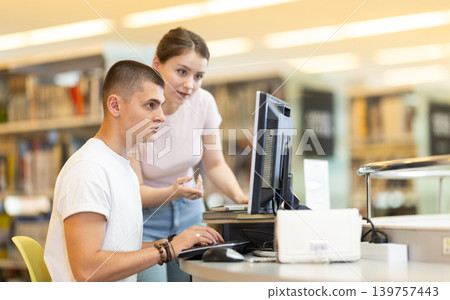 Male student works on computer in library and talks with female friend. 139757443