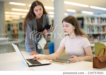 Two girls studying for an exam together in university library using books and laptop Two girls studying for an exam together in university library using books and laptop 139757693