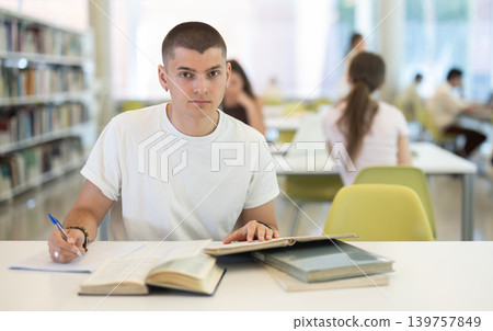 Male student sits at table in library and studies, reads books and takes notes Male student sits at table in library and studies, reads books and takes notes 139757849