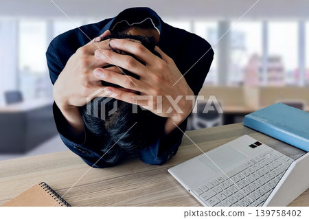 A man holding his head in his hands at his desk (office background) A man holding his head in his hands at his desk (office background) 139758402