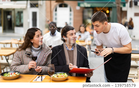 On open area of cafe, guy waiter communicates with couple of customer, accepts order On open area of cafe, guy waiter communicates with couple of customer, accepts order 139758579
