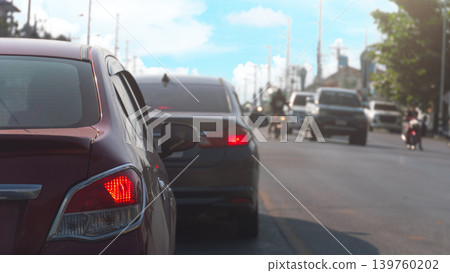 Rear angle shot focus on a red hatchback stuck in traffic on a city street. Other vehicles and motorcycle are visible ahead on the road under a clear sky highlighting daily urban commuting challenges. Rear angle shot focus on a red hatchback stuck in traffic on a city street. Other vehicles and motorcycle are visible ahead on the road under a clear sky highlighting daily urban commuting challenges. 139760202