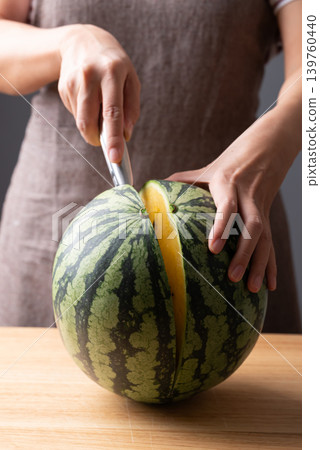 Woman's hands holding a knife and cutting a fresh yellow watermelon on a wooden cutting board Woman's hands holding a knife and cutting a fresh yellow watermelon on a wooden cutting board 139760440