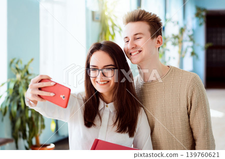 Couple of young cheerful students making selfie on red mobile phone standing indoor in university campus holding books. Happy students having good time together after classes Couple of young cheerful students making selfie on red mobile phone standing indoor in university campus holding books. Happy students having good time together after classes 139760621