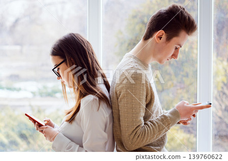 Couple of young students sitting next to one another and using mobile phones having no expression, concerned to their phones, have no interest in one another, problem of constant using modern gadgets Couple of young students sitting next to one another and using mobile phones having no expression, concerned to their phones, have no interest in one another, problem of constant using modern gadgets 139760622