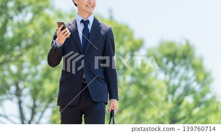 一位男性商務人士手持智慧型手機，在戶外行走。 139760714