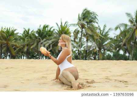 Woman sits on sand holding a seashell at the beach Woman sits on sand holding a seashell at the beach 139763096