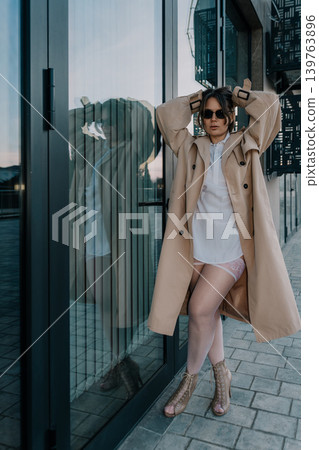Woman trenchcoat urban stylish model posing in a beige coat and sunglasses by a reflective glass building entrance Woman trenchcoat urban stylish model posing in a beige coat and sunglasses by a reflective glass building entrance 139763896