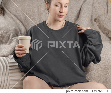Blank dark grey oversized sweatshirt mockup on fair-haired young woman sitting on sofa, front view Blank dark grey oversized sweatshirt mockup on fair-haired young woman sitting on sofa, front view 139764301