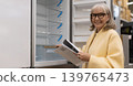 Smiling senior woman examining an empty refrigerator display in a retail store 139765473