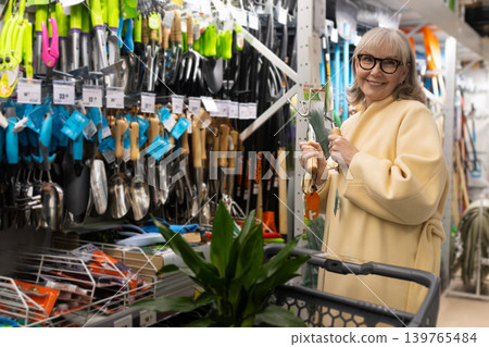 Senior woman smiling, holding gardening tools, standing by shelves in a hardware store Senior woman smiling, holding gardening tools, standing by shelves in a hardware store 139765484