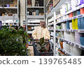 Senior woman pushing a shopping cart with a plant, looking at colorful pots on shelves 139765485