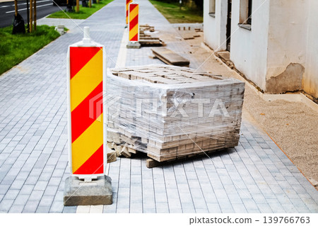 A wrapped pallet of paving stones and red and yellow safety barriers sit on a partially finished gray sidewalk during an urban street construction project. 139766763