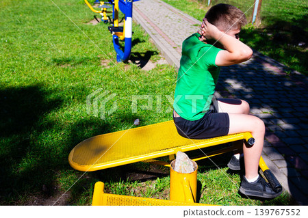 Boy exercising outdoors on yellow sit-up bench. Healthy lifestyle, fitness, and active kids concept. Boy exercising outdoors on yellow sit-up bench. Healthy lifestyle, fitness, and active kids concept. 139767552